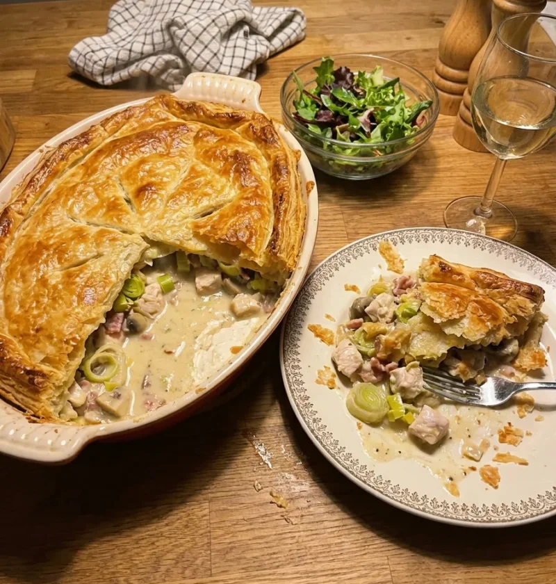 Gordon Ramsay chicken pie in an oval dish with golden puff pastry lid, a wedge cut out showing creamy sauce with chicken leeks bacon and mushrooms, served on a plate beside it