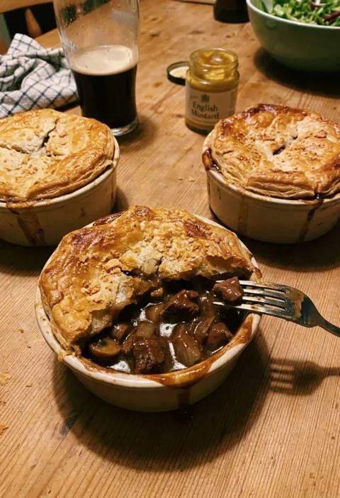 Gordon Ramsay steak and ale pie in individual dishes with golden puff pastry lids, one broken open showing dark Guinness beef and mushroom filling, a pint of dark beer and English mustard on the table
