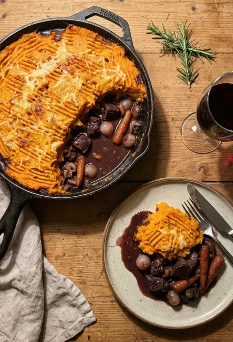 Gordon Ramsay venison pie in a cast-iron pan with orange sweet potato mash topping, a portion scooped out showing dark venison filling with baby onions carrots and mushrooms in wine sauce