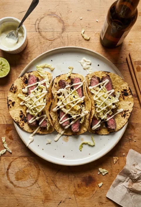 Three beef tacos in charred corn tortilla shells with pink medium-rare sirloin slices pale pickled Chinese cabbage and wasabi mayo drizzle on a plate