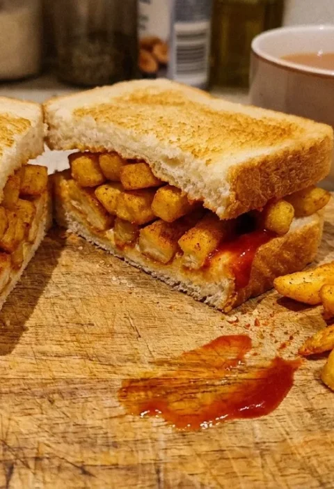Gordon Ramsay chip butty sliced in half on a wooden chopping board showing oven chips with smoked paprika and ketchup inside crusty white bread with loose chips and a mug of tea