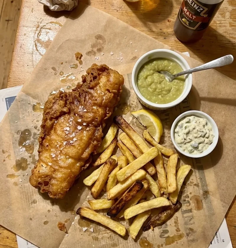 Golden beer-battered cod with thick-cut chips mushy peas and tartar sauce on greaseproof paper with a pint of lager and malt vinegar