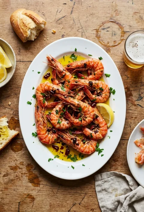 Shell-on garlic prawns in golden olive oil with chopped garlic dried chilli and parsley on a white plate with lemon wedges and crusty bread