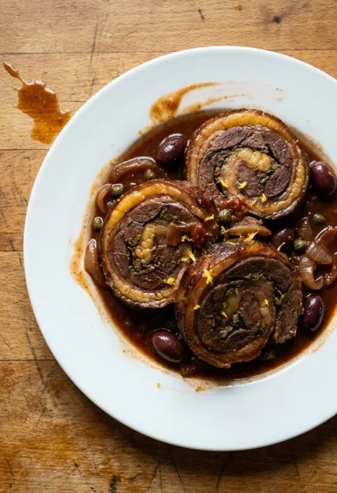 Gordon Ramsay stuffed lamb breast sliced into thick spirals in a rich tomato sauce with black olives capers and braised onion topped with lemon zest on a white plate