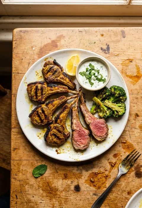 Gordon Ramsay aromatic lamb chops with turmeric spice rub griddle marks and pink medium rare interior served with minty yoghurt chargrilled broccoli and lemon on a white plate