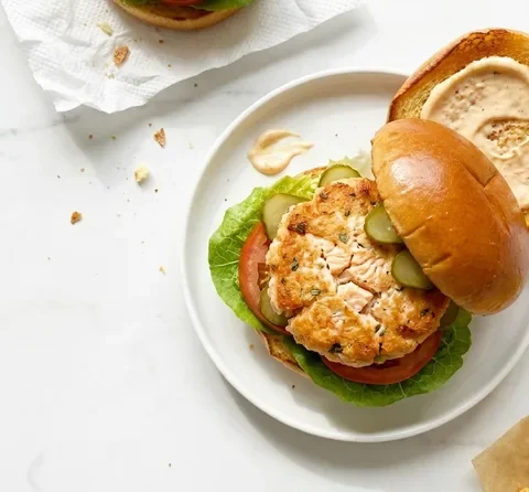 Gordon Ramsay salmon burger with golden salmon patty, lettuce, tomato and gherkins in a toasted brioche bun with sriracha mayo on the lifted lid