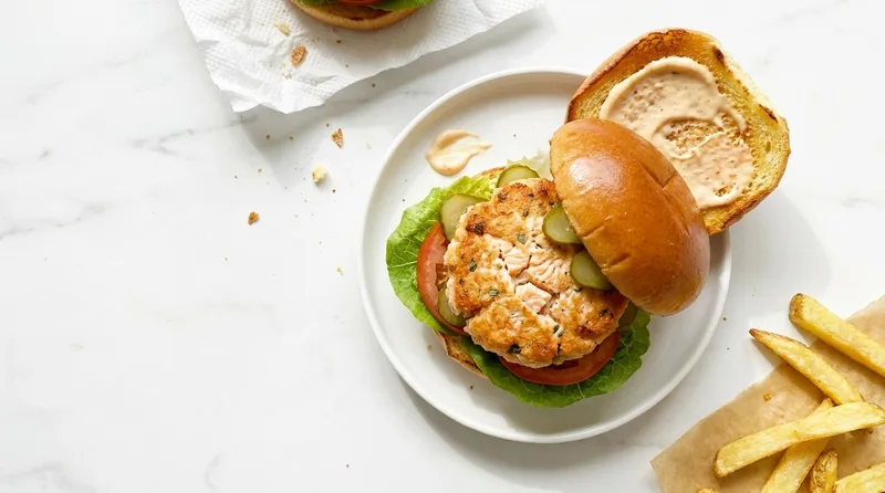 Gordon Ramsay salmon burger with golden salmon patty, lettuce, tomato and gherkins in a toasted brioche bun with sriracha mayo on the lifted lid