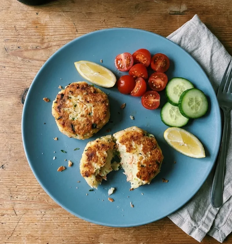 Gordon Ramsay salmon fish cakes coated in golden breadcrumbs, one broken open showing potato and salmon, with cherry tomatoes and cucumber