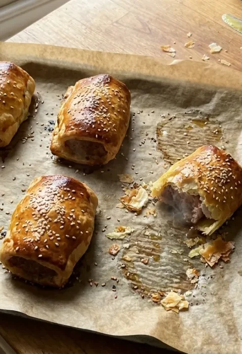 Gordon Ramsay sausage rolls fresh from the oven: six golden puff pastry rolls with sesame seeds on greaseproof paper, one broken open showing the sausage filling, with English mustard and pastry brush