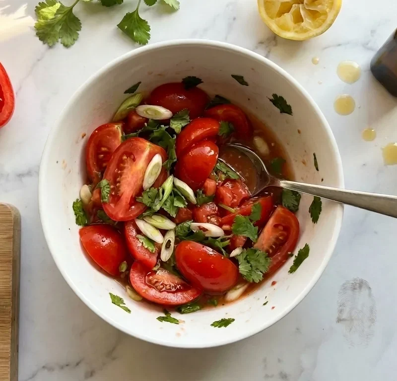 Gordon Ramsay tomato salsa: chunky halved plum tomatoes with diagonal spring onion slices and coriander in a white bowl, with sesame oil bottle, hot sauce and squeezed lemon