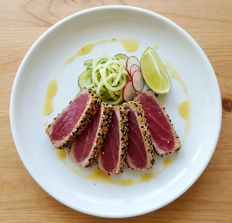 Gordon Ramsay sesame crusted tuna steak sliced showing ruby pink rare centre with black and white sesame seed crust pickled cucumber noodles radish slices lime wedge and yuzu dressing on a white plate