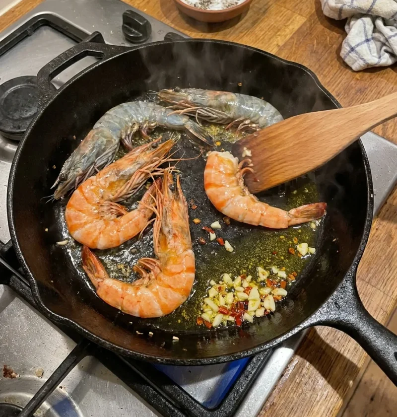 Shell-on tiger prawns searing in a cast iron pan with garlic and chilli on a gas hob
