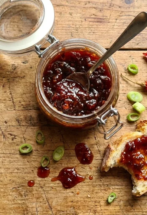 Gordon Ramsay chilli jam in a glass jar with a spoon red chillies and spring onions on a wood table